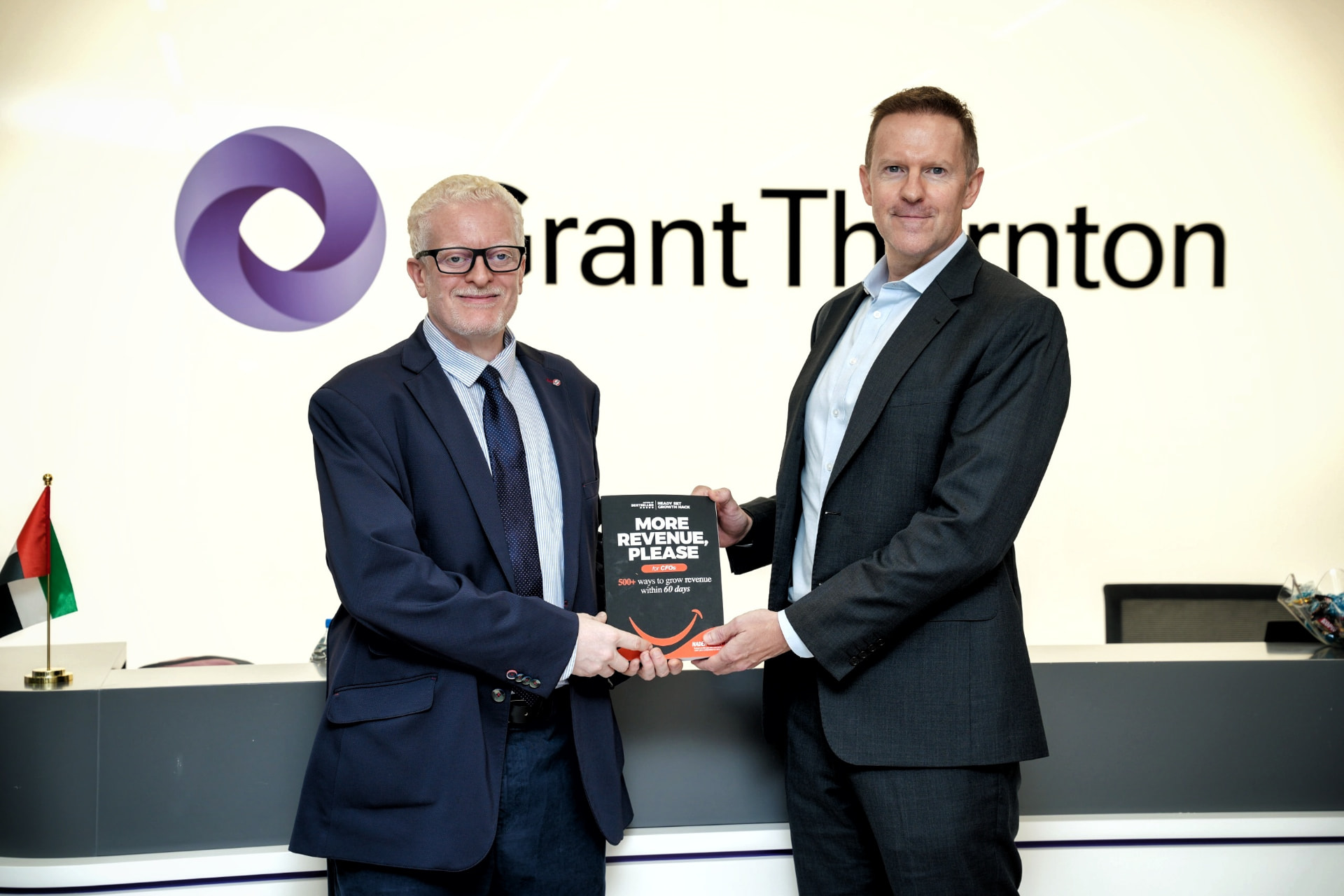 Nader Sabry holding a copy of More Revenue, Please with a colleague in front of the Grant Thornton logo at Grant Thornton UAE